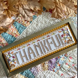 May include: A framed cross-stitch piece with the word "THANKFUL" in tan thread. The frame is a muted green, and the background is a patchwork quilt. The text "Stitching with the Housewives" and "Word Swap" are also visible.
