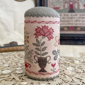 May include: A cylindrical pin cushion featuring a light pink fabric base with cross-stitched floral motifs in shades of red, brown, and green. The top and bottom edges are finished with a grey scalloped trim. The text Summer Garden Drum is visible.