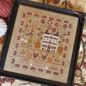 May include: A framed cross-stitch sampler featuring a house, a scarecrow, pumpkins, and floral borders on a tan fabric background. The design includes an alphabet, the word AUTUMN, and various seasonal motifs in shades of red, orange, and brown.
