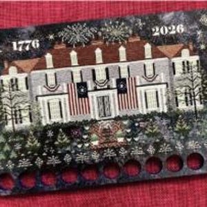 May include: A decorative wooden needle minder with a painted image of a stately building, American flags, and the years 1776 and 2026. The needle minder has a dark background with a starry design and a series of holes along the bottom. A smaller matching pin is included.