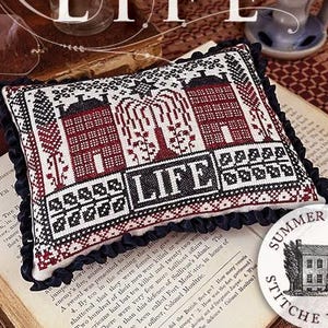 May include: A decorative pillow with a cross-stitch design featuring two red houses, a weeping willow, and the word "LIFE." The pillow has a black ruffled edge and is set on an open book. The text "Number One DECLARATION COVERLETS LIFE" is also visible.