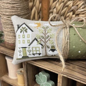 May include: A display of handcrafted items, including a cross-stitched pillow with a house design, a green polka-dot fabric roll, and a black pot of gold. The display is set in a wooden organizer with spools, a horseshoe, and a shamrock. Text reads "EWE HAPPY HOME" and "PRIMROSE COTTAGE."