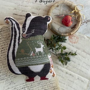 May include: A handmade skunk plushie from the "Animal Cracker Series + Sugar+" collection. The skunk wears a green sweater with a deer design. The plushie has black and white stripes and red boots. The design is by Stacy Nash.