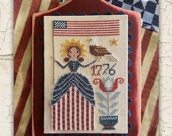 May include: A patriotic cross-stitch design featuring an American flag, an eagle, and a woman in a red, white, and blue dress. The design is mounted on a dark red wooden plaque with a handle. The background includes striped and checkered fabric.