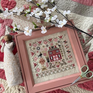 May include: A framed cross-stitch embroidery featuring a red house with a white picket fence and a garden with flowers and birds. The frame is a light pink color and the embroidery is on a cream-colored fabric.