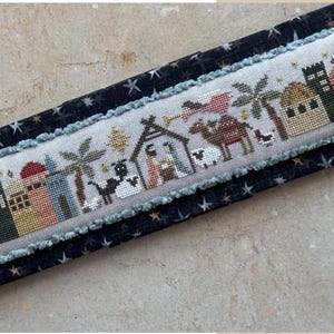 May include: A cross-stitch pattern depicting the nativity scene with a star, a camel, a shepherd, a donkey, a sheep, a barn, and a city skyline. The pattern is stitched on a white fabric with a blue border.