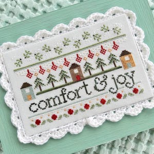 May include: Cross-stitch embroidery with the words "comfort & joy" in black. The design includes houses, trees, and red ornaments. The embroidery is framed with a white, scalloped lace border and set on a light green background.