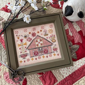 May include: A framed cross-stitch design of a red barn with a white door and windows. The barn is surrounded by flowers and other details. The frame is a dark green color and is sitting on a red and white quilt.