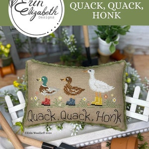 May include: Cross stitch pattern featuring three ducks and a goose with the text "Quack, Quack, Honk" on a green pillow. The pillow is sitting on a white tray with gardening tools.