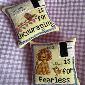 May include: Two small, square, cross-stitched pillows. One features a bird on a branch with the text "Fly High Little One" and "is for Encouraging". The other has a lion and a chick with the text "Boo! LOL! is for Fearless".