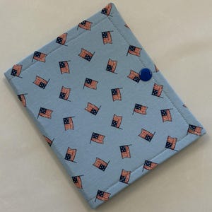 May include: A light blue fabric item with a repeating pattern of American flags in red, white, and blue. The item is rectangular with rounded corners and a blue button closure. The edges are stitched with a contrasting thread.