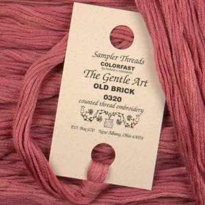 May include: Close-up of a skein of Old Brick colored embroidery thread with a coordinating tag. The tag reads "Sampler Threads, The Gentle Art, 0320, counted thread embroidery." The thread is a muted reddish-brown color, perfect for cross-stitch and embroidery projects.