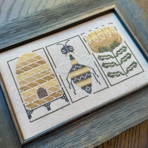 May include: A framed cross-stitch embroidery featuring a bee, a beehive, and a flower. The bee is yellow and black with a striped pattern. The beehive is brown and yellow with a honeycomb pattern. The flower is yellow with green leaves.