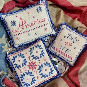 May include: Three patriotic, embroidered pillows with blue ruffled edges. One pillow reads "America" in red script, another says "July 4th 1776", and the third displays the numbers "2017". The pillows are on a silver tray.