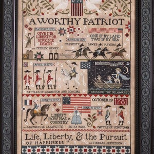 May include: A framed cross-stitch sampler with a patriotic theme. The artwork features historical scenes, dates, and text including "A Worthy Patriot" and "Life, Liberty, & the Pursuit of Happiness." The color palette includes beige, red, blue, and green.