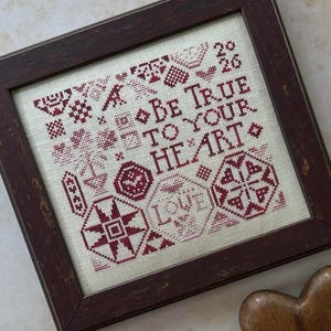 May include: A framed cross-stitch sampler with the text "Be True To Your Heart" in burgundy thread. The design includes geometric shapes, hearts, and floral motifs. The frame is a dark, stained wood.