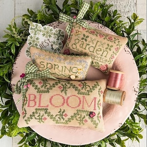May include: A collection of embroidered spring-themed cartouches, featuring the words "SPRING" and "BLOOM", along with a "GARDEN" design. The pieces are adorned with floral patterns, green accents, and a ladybug. The image includes spools of thread and greenery.