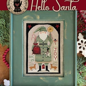 May include: A cross stitch pattern featuring a green-clad Santa Claus holding a red bag and a sign that says "JOY". The pattern is framed in a green frame with a distressed finish. The title "Hello Santa" is written in red cursive above the frame. The text "7 Cross Stitch Charts by Teresa Kogut" is written below the frame.