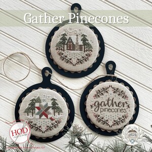 May include: Three round cross-stitch ornaments with a rustic theme. The ornaments feature a cabin in the woods, a patchwork quilt, and the text "gather pinecones".