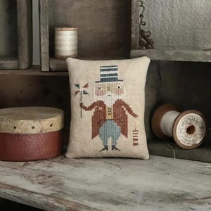 May include: A small, beige pillow with a cross-stitch design of a man in a red coat and a tall hat holding a flag. The pillow is sitting on a wooden shelf with spools of thread and a wooden box.