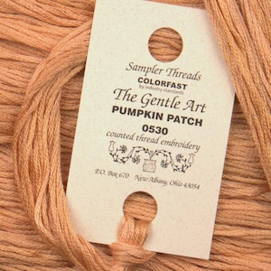 May include: A close-up of a skein of orange embroidery floss with a white label that reads "Sampler Threads COLORFAST by industry standards The Gentle Art PUMPKIN PATCH 0530 counted thread embroidery P.O. Box 670 New Albany, Ohio 43054"