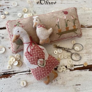 May include: A handcrafted textile art piece featuring a large goose and a smaller duck, both with detailed cross-stitch patterns in red and white. The design includes the text "Animal Cracker Series + Olive+" and "Design by Stacy Nash".