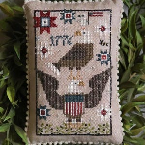 May include: A rectangular, beige, cross-stitched pillow featuring an American eagle design. The eagle is brown and white, with a red, white, and blue shield. The year "1776" is stitched above the eagle. The pillow is trimmed with a white pom-pom border.