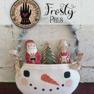 May include: A handmade snowman basket with a silver tinsel handle, holding embroidered Christmas figures: Santa Claus, a Christmas tree, and a woman. The basket has a carrot nose and black dot eyes. The text "Frosty Pals" is above the basket.