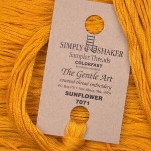 May include: A brown label with the text "SIMPLY SHAKER Sampler Threads COLORFAST by industry standards The Gentle Art counted thread embroidery PO Box 670 New Albany, Ohio 45054 SUNFLOWER 7071" on a spool of yellow embroidery floss.