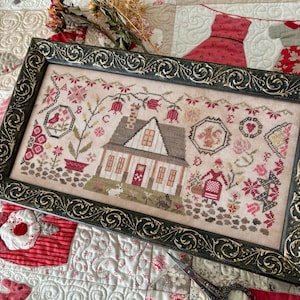 May include: Framed cross-stitch artwork featuring a detailed depiction of a house with a brown roof, surrounded by floral and geometric patterns. The frame is dark with ornate detailing. The artwork is on a quilted background.