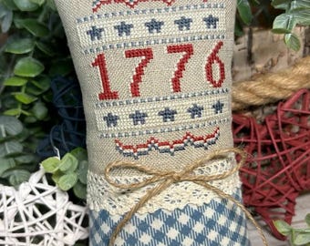 Puede incluir: Un cojín decorativo con tela de lino beige, con bordado de punto de cruz del número "1776" en rojo. La parte superior tiene detalles en rojo y azul, y la parte inferior tiene un patrón de cuadros azules y blancos. Un ribete de encaje y un lazo de cuerda añaden detalles.