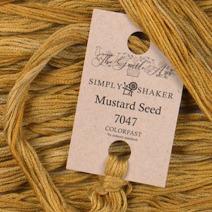 May include: Mustard Seed embroidery floss from The Gentle Art. The label reads "SIMPLY SHAKER Mustard Seed 7047 COLORFAST". The floss is a golden yellow color, with a slight variation in tone. The floss is bundled together.