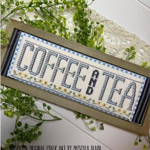 May include: A framed cross-stitch sign with the words "COFFEE & TEA" in gray and black lettering. The sign has a light blue and yellow floral border and is set in a wooden frame. The text "BASED ON ORIGINAL CHALK ART BY PRISCILLA BLAIN" is at the bottom.