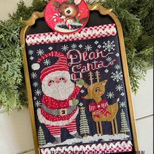 May include: A framed Christmas cross-stitch featuring Santa Claus and a reindeer, with the words "Dear Santa" embroidered on a black background. The frame is gold, and a reindeer ornament hangs at the top. The design includes snowflakes and Christmas trees.