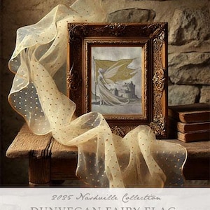 May include: A framed cross-stitch pattern featuring a fairy holding a flag with the text "Dunvegan Fairy Flag". The frame is gold and the background is a rustic stone wall.