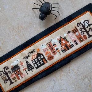 May include: A cross-stitch pattern featuring a row of houses with orange roofs and white trim, a black cat, and a spooky tree. The houses are decorated with Halloween-themed details, such as ghosts, bats, and pumpkins. The pattern is stitched on a cream-colored fabric.