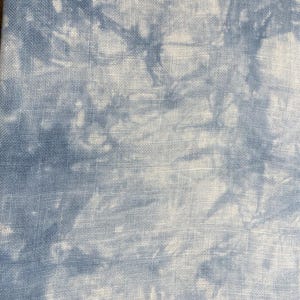 May include: Close-up of a fabric with a blue and white tie-dye pattern. The fabric has a textured appearance, with the blue dye creating a cloudy effect against the white background. The fabric is likely cotton or linen.