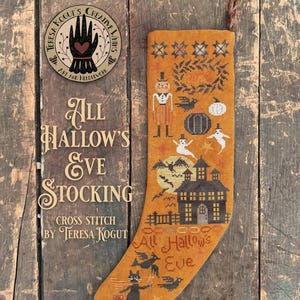 May include: A mustard-colored fabric stocking featuring a cross-stitch design with Halloween motifs including a pumpkin-headed figure, ghosts, a haunted house, a black cat, bats, and the text "All Hallow's Eve" with a braided hanging loop at the top.