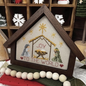 May include: A wooden framed cross-stitch depicting a nativity scene. The cross-stitch features a stable with the word "JOY" and the phrase "TO THE WORLD". The frame is dark brown, and the cross-stitch has gold, blue, and green accents.