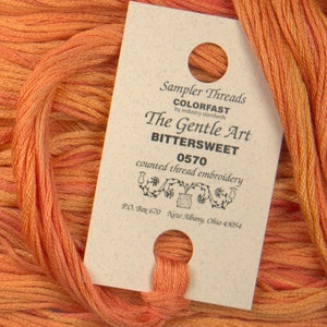 May include: A close-up of a spool of orange embroidery floss with a label that reads "Sampler Threads COLORFAST by industry standards The Gentle Art BITTERSWEET 0570 counted thread embroidery P.O. Box 670 New Albany, Ohio 43054"
