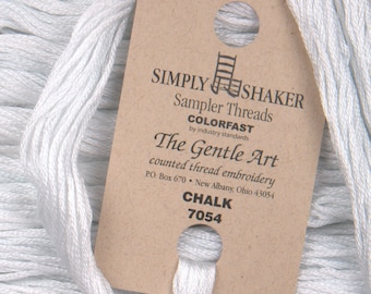 May include: Close-up of white embroidery thread with a brown tag. The tag reads "SIMPLY SHAKER Sampler Threads COLORFAST The Gentle Art counted thread embroidery CHALK 7054". The thread is a light, neutral color.