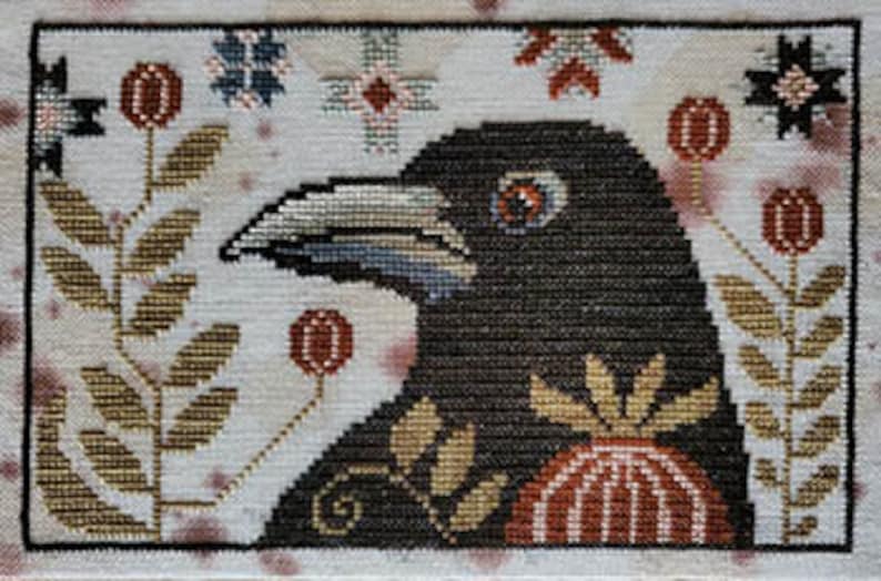 Op de afbeelding: Borduurpatroon met een zwarte raaf met een rood oog, omringd door gestileerde bloemen en bladeren in tinten bruin, rood en blauw op een witte achtergrond.