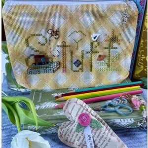 May include: A zippered pouch with a yellow and white checkered pattern featuring green cross-stitched text that reads "stuff" with embroidered book, pencil, and flower motifs. A silver charm is attached to the zipper pull of the fabric pouch.