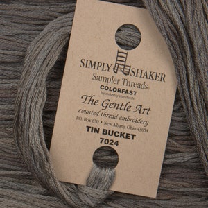 May include: A skein of gray embroidery floss with a label that reads "SIMPLY SHAKER Sampler Threads COLORFAST by industry standards The Gentle Art counted thread embroidery P.O. Box 670 New Albany, Ohio 43054 TIN BUCKET 7024"