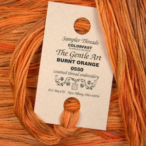 May include: A close-up of a spool of burnt orange embroidery floss with a label that reads "Sampler Threads Colorfast by industry standards The Gentle Art Burnt Orange 0550 counted thread embroidery P.O. Box 670 New Albany, Ohio 43054"