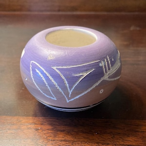 May include: A round, hand-painted ceramic bowl with a wide opening. The bowl is primarily lavender with a gradient to tan at the base. White leaf-like designs are etched into the surface. A black and white stripe encircles the base.