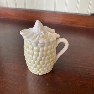 May include: A ceramic salt shaker shaped like an ear of corn, with a handle and a lid. The shaker is light yellow with a textured surface, and the lid is white. The handle is small and curved.
