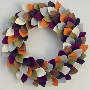 May include: A circular wreath constructed from layered, petal-shaped felt pieces in a palette of deep purple, burnt orange, olive green, brown, cream, and light gray, arranged in an overlapping pattern to create a textured, multi-colored display.