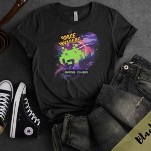 May include: Black t-shirt featuring a Space Invaders graphic with a green pixelated alien, a purple planet, and the words "GAME OVER." The shirt is paired with black high-top sneakers, dark wash jeans, and a brown belt.