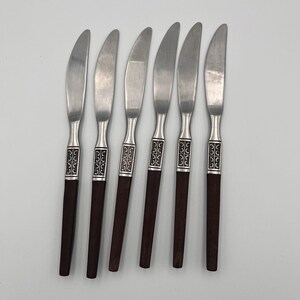 May include: Set of six stainless steel knives with dark brown wooden handles. The handles have a decorative silver band with a repeating geometric pattern. The blades are curved and have a polished finish. Ideal for a formal dining setting.